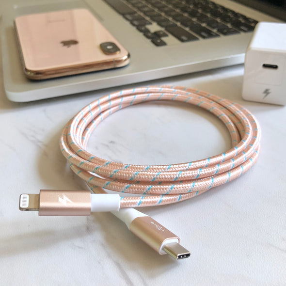 Braided lightning to USB-C charging cable in a pink and white pattern placed on a white surface. In the background, there is a pink smartphone, a laptop with a partially visible keyboard, and a white charger block with a stylized lightning bolt icon.