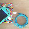 The ChargeCords Minty Fresh Solar Bank, boasting four blue LED indicators and quick charging capabilities, is connected to a device using a blue and yellow patterned charging cable. The turquoise solar power bank lies on a colorful, geometrically patterned surface, with a wooden tabletop in the background.