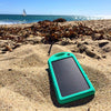 The Minty Fresh Solar Bank from ChargeCords, a rapid-charging solar-powered battery charger encased in turquoise, rests on a sandy beach amidst scattered seaweed. The serene backdrop of a calm ocean and clear blue sky, punctuated by a distant sailboat, highlights its suitability as the perfect portable external battery for beachgoers.