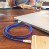 A coiled ChargeCords Varsity Lightning Cable, blue and red in color, is placed on a wooden table. It connects a silver laptop to a gold iPhone while featuring tangle-proof fabric to ensure durability and convenience. In the background, a person enjoys reading a book with a cup of coffee nearby. This Apple MFi Certified charging cable measures 5 feet (1.5 meters) in length.